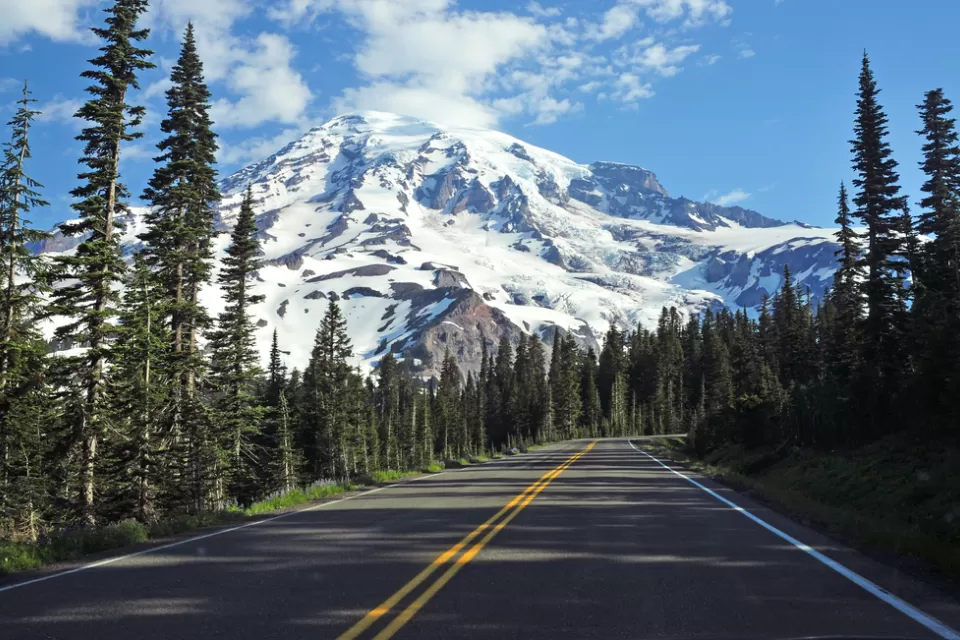 Two-lane asphalt road in Washington state with pine trees along sides and Mt. Rainier in background